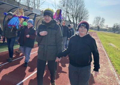 Dwoje dzieci trzymających się za ręce spaceruje po bieżni, w tle grupa osób niesie dekoracje na patykach. Słoneczny dzień.