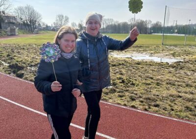 Dwie kobiety idą po bieżni sportowej, każda trzyma w rękach ozdobne kwiatki. W tle widać mur i boisko.