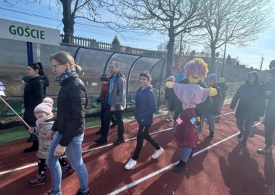 Grupa osób, w tym kobiety i dziecko, spaceruje po bieżni sportowej, niosąc kukłę Marzanny. W tle widać drzewa i mur.