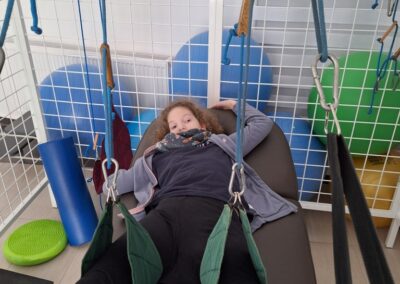 Dziecko leży na łóżku terapeutycznym, przypięte do pasów w ramach terapii zawieszeniowej, z tyłu widoczne kolorowe piłki rehabilitacyjne.