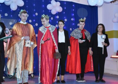 Trzej mężczyźni przebrani za królów w czerwonych pelerynach i koronach stoją na scenie. Obok nich opiekunowie.
