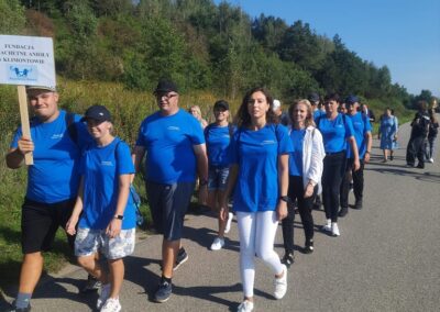 Grupa osób ubrana w niebieskie koszulki idzie drogą, prowadząc marsz. Jeden z uczestników trzyma tabliczkę z napisem Fundacja Szlachetne Anioły w Klimontowie. W tle widać drzewa i niebieskie niebo.