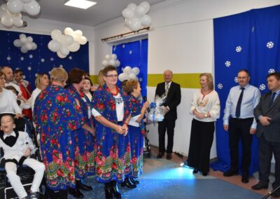 Kilka osób w kolorowych strojach ludowych stoi na scenie. Obok nich osoby w eleganckich ubraniach. W tle dekoracje z balonów i śnieżynki.