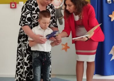 Dwie kobiety pomagają chłopcu z książką, scena w pomieszczeniu z dziecięcymi rysunkami.
