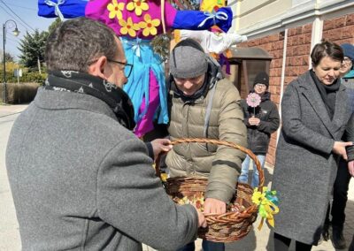 Mężczyzna wrzuca słodycze do koszyka, który trzyma dziecko na wózku inwalidzkim. W tle grupa osób.
