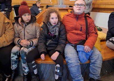 Trójka dzieci siedząca na ławce w drewnianym wnętrzu, uczestnicząca w spotkaniu świątecznym.