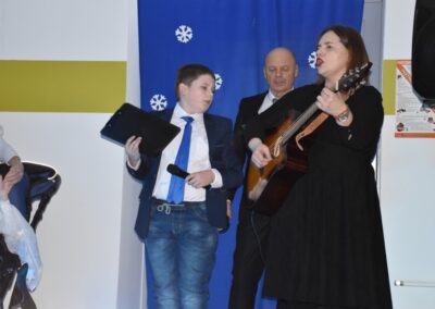 Kobieta grająca na gitarze śpiewa, a obok niej stoi chłopiec z mikrofonem i tabletem. W tle dekoracje ze śnieżynkami.