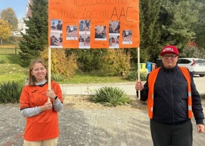 Dwie osoby z pomarańczowym plakatem AAC na zewnątrz, w tle drzewa i budynek.