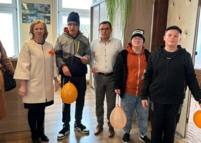 Pięć osób w pokoju z drewnianą podłogą, rośliną doniczkową i trzema pomarańczowymi balonami.
