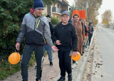 Grupa ludzi z pomarańczowymi balonami idzie chodnikiem w jesiennych ubraniach, w tle drzewa.