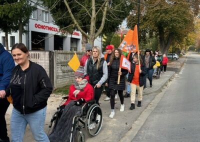 Grupa na chodniku obok budynku straży pożarnej, osoba na wózku, pomarańczowe flagi.