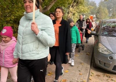 Grupa ludzi idąca chodnikiem obok zaparkowanych samochodów, jedna osoba niesie plakat.