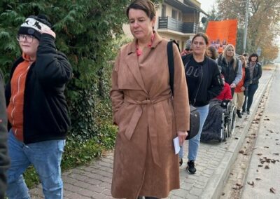 Grupa ludzi maszeruje chodnikiem wzdłuż ulicy, prowadząca osoba w długim płaszczu.