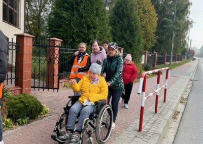 Grupa ludzi idzie chodnikiem, jedna osoba na wózku inwalidzkim. W tle drzewa i ogrodzenie.
