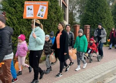 Grupa ludzi idzie chodnikiem, w tle budynek i drzewa, uczestnik trzyma tablicę informacyjną.