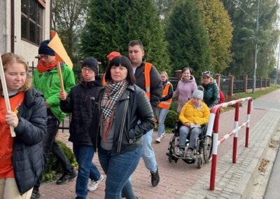 Grupa ludzi na chodniku, jedna osoba na wózku, odblaskowe kamizelki, pomarańczowa flaga, drzewa.
