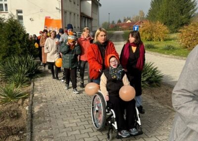 Grupa ludzi na chodniku, w tym osoba na wózku, z balonami. W tle budynki i roślinność.