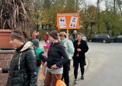 Grupa maszeruje ulicą z tablicą informacyjną i pomarańczowymi balonami; w tle drzewa, auta.