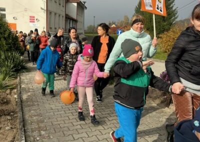 Grupa osób, w tym dzieci z balonami, spaceruje chodnikiem obok budynku w pochmurny dzień.
