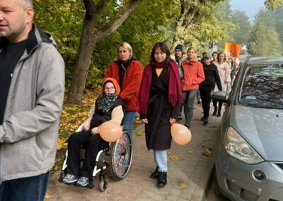 Grupa ludzi spaceruje chodnikiem w parku jesienią, osoba na wózku z balonami, auta obok.