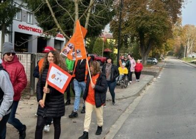 Ludzie na chodniku z banerami, budynek Ochotniczej Straży Pożarnej w tle, drzewa po bokach.