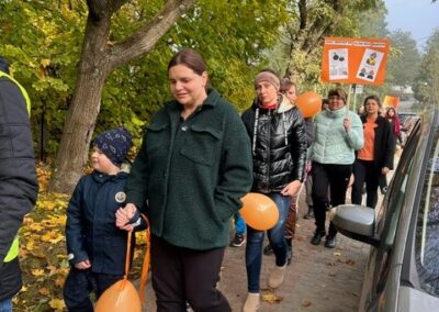 Ludzie spacerują jesiennym chodnikiem z pomarańczowymi balonami i plakatem w tle.