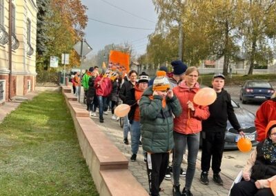 Ludzie maszerują z pomarańczowymi balonami i transparentem obok budynku w jesiennym otoczeniu.