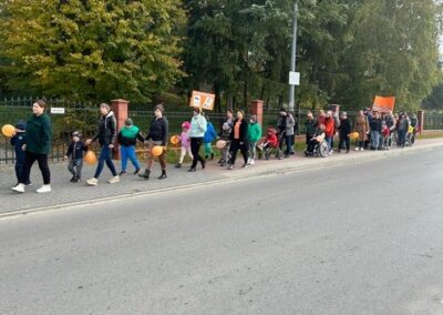 Grupa ludzi maszeruje chodnikiem z pomarańczowymi balonami i plakatami, tło stanowią drzewa.