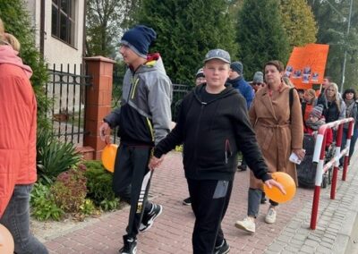 Ludzie na chodniku z pomarańczowymi balonami, drzewa i krzewy w tle.