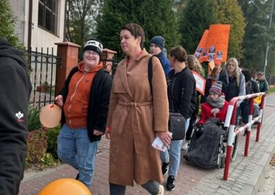 Grupa ludzi spaceruje chodnikiem z pomarańczowymi balonami i transparentami; drzewa w tle.