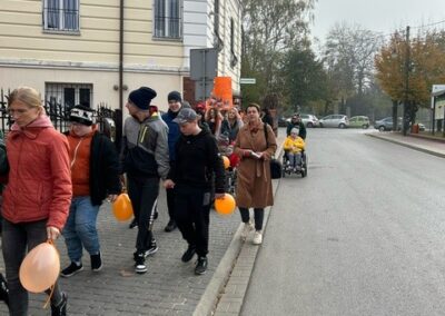 Grupa osób idzie chodnikiem z pomarańczowymi balonami, w tle budynek i drzewa.