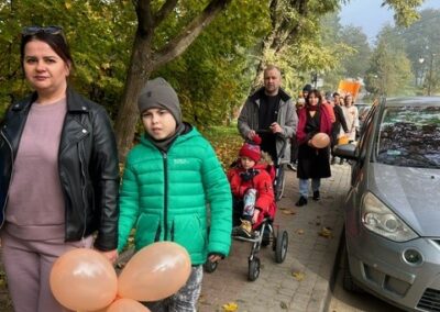 Grupa ludzi spacerująca chodnikiem wśród jesiennych drzew, dwie osoby z pomarańczowymi balonami.