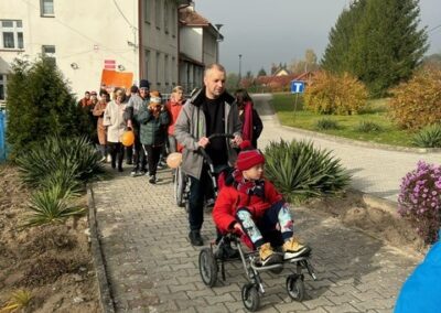 Grupa ludzi idąca na zewnątrz, prowadzi osobę z wózkiem, w tle budynek i zieleń.
