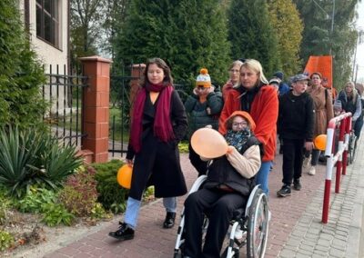 Grupa ludzi idzie chodnikiem, osoba na wózku inwalidzkim trzyma pomarańczowy balon.