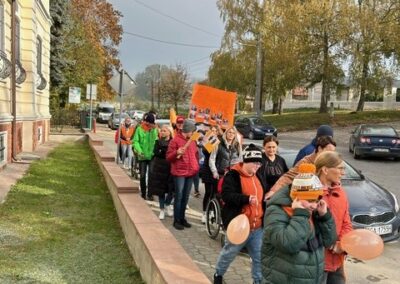Ludzie maszerują z pomarańczowymi balonami i tablicą obok budynku i samochodów.