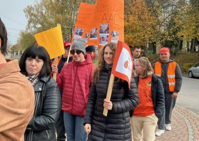 Ludzie maszerują z pomarańczowymi i żółtymi transparentami na tle jesiennych drzew.