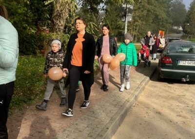 Grupa ludzi z dziećmi spaceruje chodnikiem, niektórzy trzymają brązowe balony.