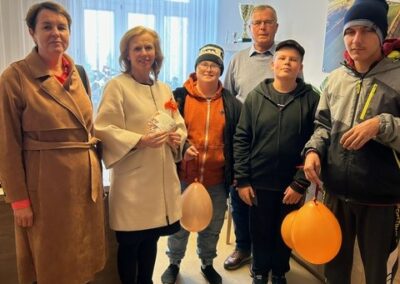 Sześć osób z balonami stoi w pokoju z trofeami na półce i dużym oknem w tle.