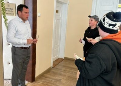 Trzech mężczyzn rozmawia w korytarzu przy jasnobeżowej ścianie z tabliczką.