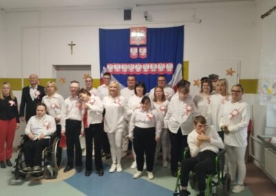 Grupa osób w białych bluzkach z kotylionami, tło z napisem "11 LISTOPADA" i godłem Polski.