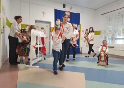 Dzieci bawią się z konikami na patyku w sali, nauczyciele pomagają, polskie dekoracje w tle.