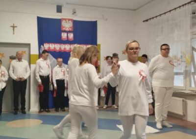 Grupa osób w białych strojach na tle niebieskiego sztandaru z orłem, ustawiona w półkole.