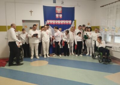 Grupa ludzi w białych strojach przed niebieskim tłem z polskimi symbolami narodowymi.