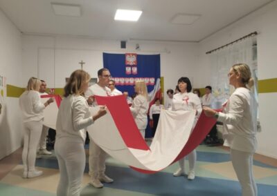 Osoby w białych ubraniach trzymają dużą flagę Polski, w tle flaga z godłem Polski.
