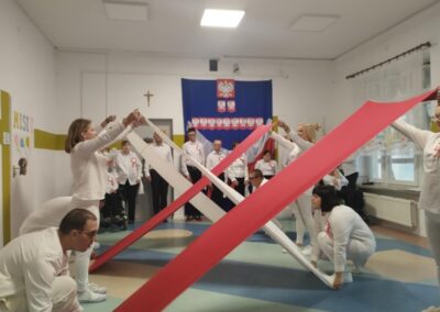 Grupa ludzi w białych strojach trzyma czerwone i białe taśmy, tworząc krzyż w sali z flagą Polski.