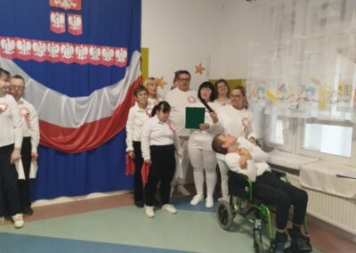 Grupa osób w białych koszulach z czerwonymi ozdobami, przed niebieskim tłem z polską flagą.