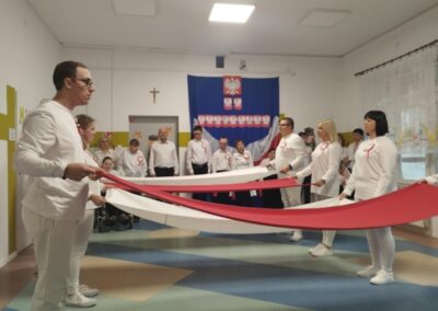 Ludzie w białych ubraniach trzymają biało-czerwony materiał na tle z polskim godłem i napisem.