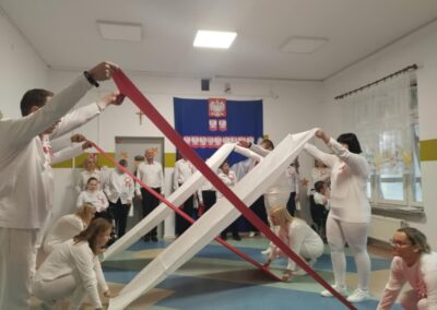 Grupa ludzi w białych strojach z biało-czerwonymi wstęgami, flaga Polski w tle.