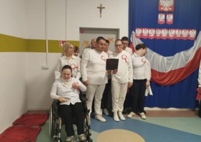 Grupa osób ubranych na biało świętuje 11 listopada przy polskiej fladze, jedna osoba na wózku.