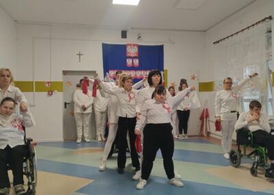 Osoby w białych strojach występują w sali z polską flagą i godłem, synchronizują ruchy.